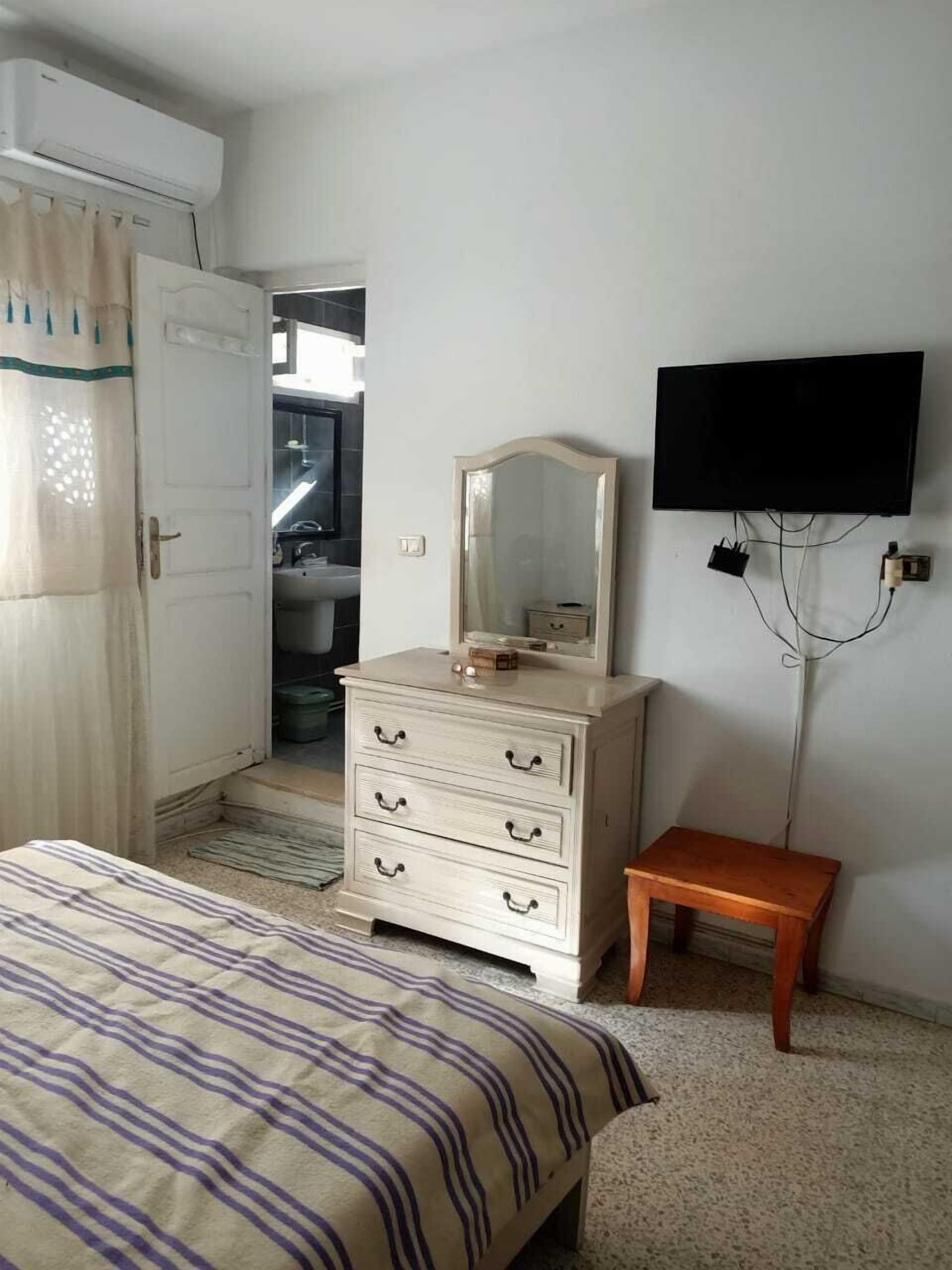 A bedroom with a bed, a dresser with drawers, a mirror, a television mounted on the wall, and a small wooden stool.