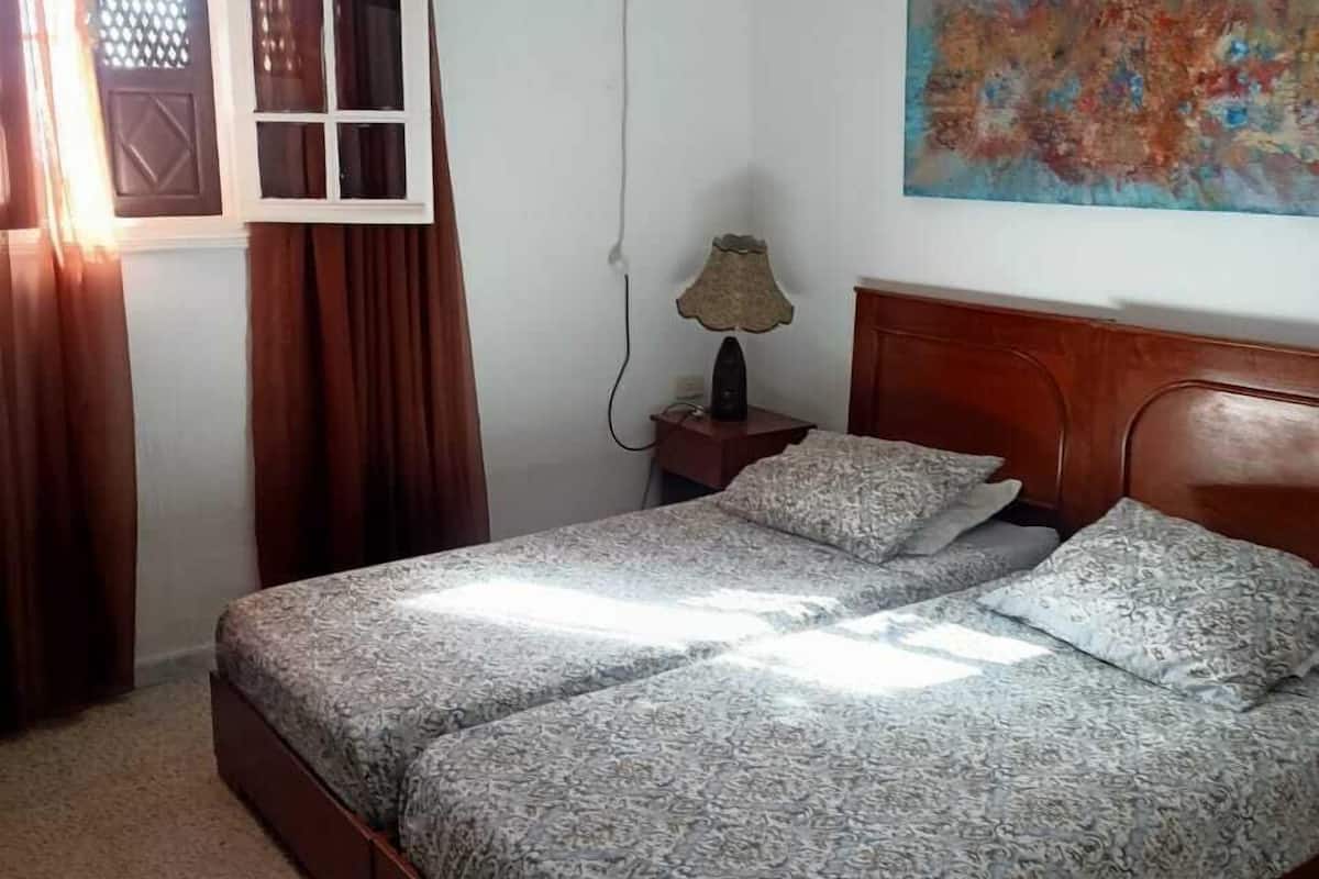 Ein Schlafzimmer mit Bett, Nachttisch, Lampe, einem Fenster mit Vorhängen und einem Wandbild.