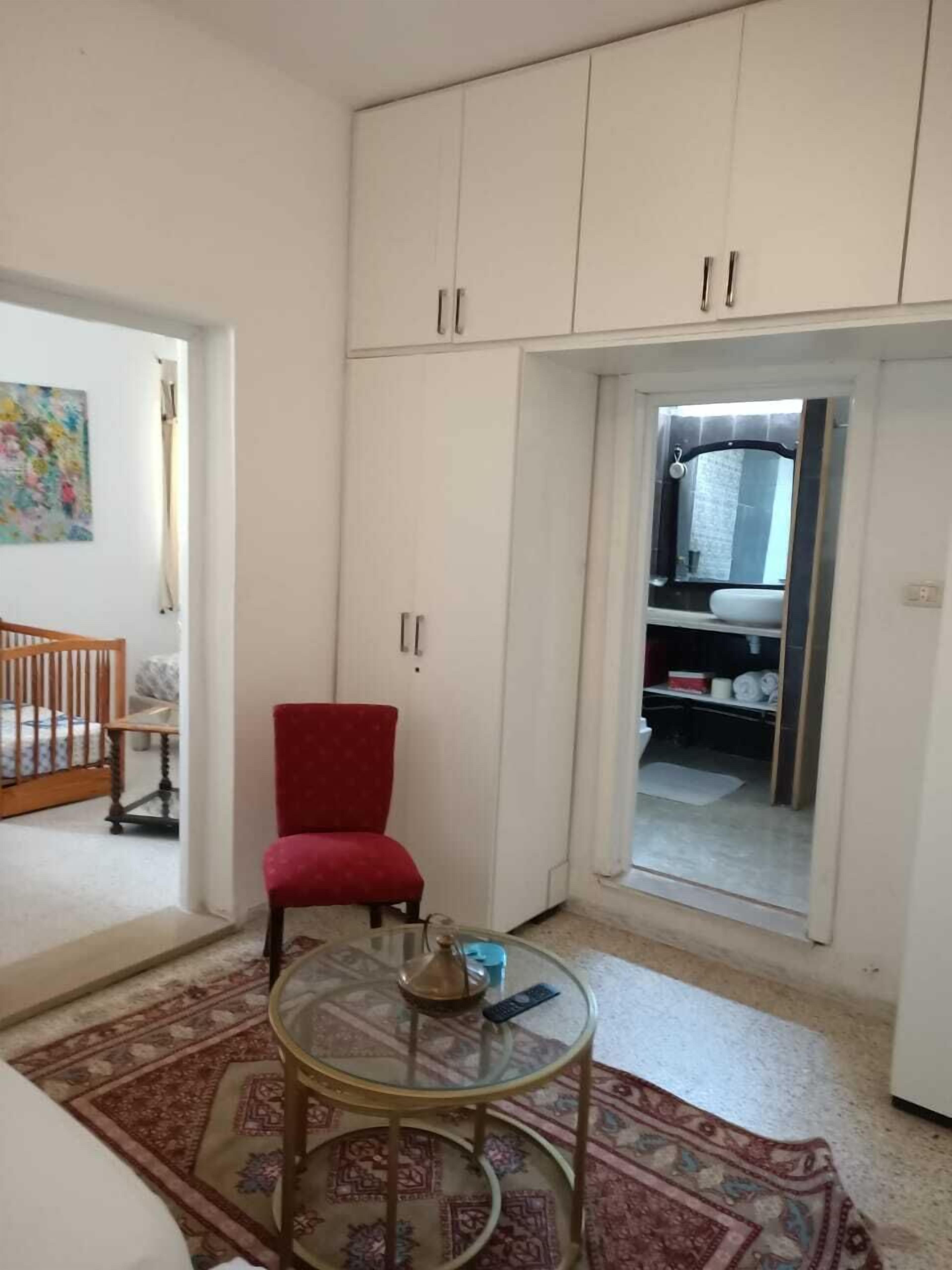 A room with a red chair, a glass table, and a patterned rug. There is a crib in the corner and a wardrobe with a mirror.