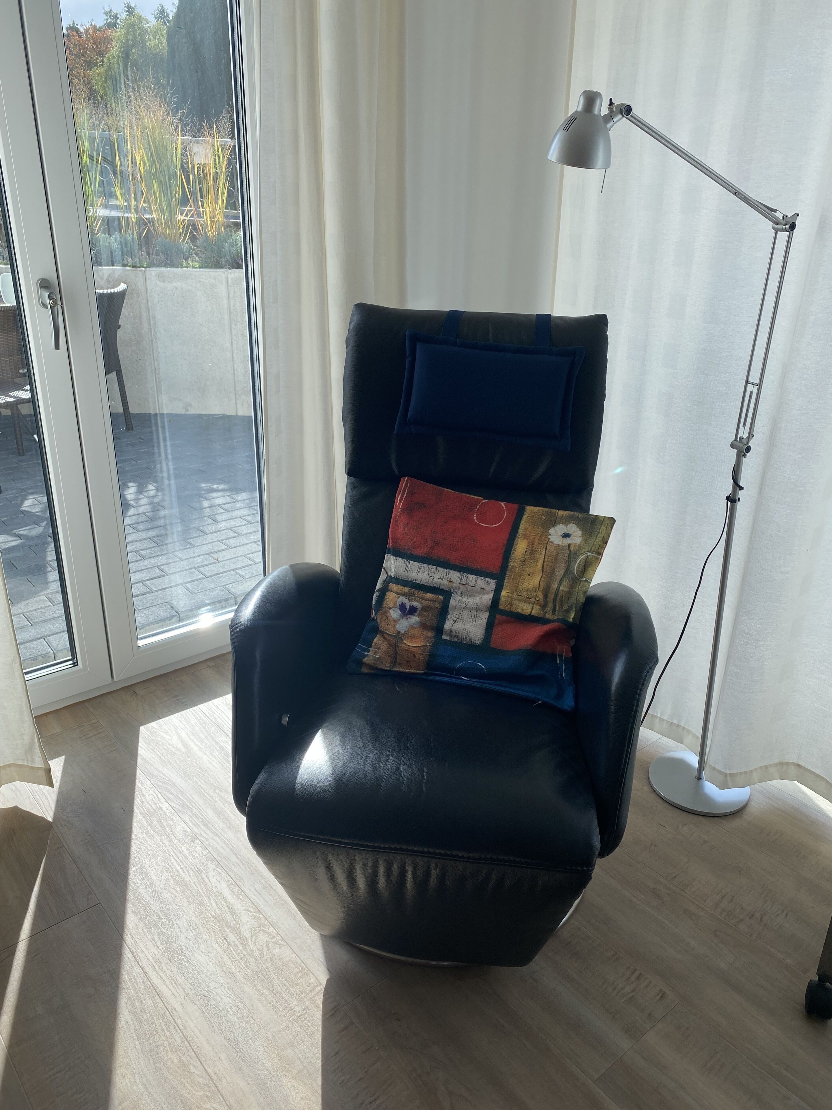 Un fauteuil en cuir noir agrémenté d’un coussin coloré, placé près d’une fenêtre donnant sur un jardin et une rue.