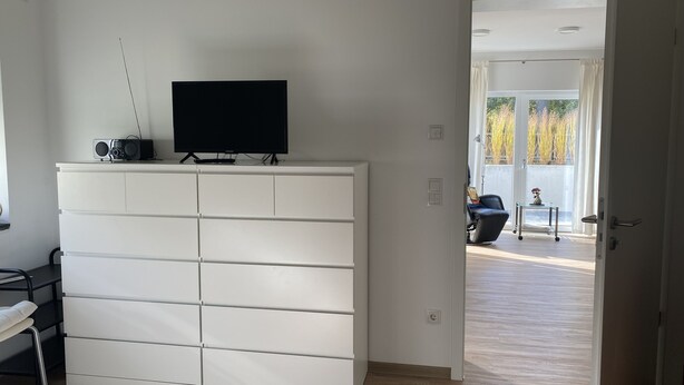 Una habitación moderna con un armario blanco, un televisor de pantalla plana y una puerta que da a un balcón con mobiliario exterior.