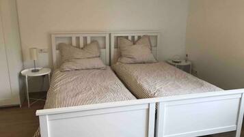 Una cama doble con estructura blanca y ropa de cama a rayas, una mesita de noche con lámpara y una pequeña mesa redonda con una botella.