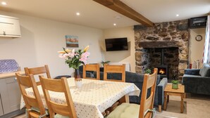 A cozy living room with a stone fireplace, a dining area with a table and chairs, and a television mounted on the wall. - Pennant Igyn (Pwllheli)