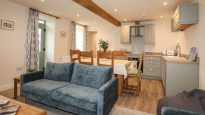 A cozy living room with a blue sofa, wooden dining table, and chairs. A kitchen with modern appliances and a small island is visible in the background. - Pennant Igyn (Pwllheli)