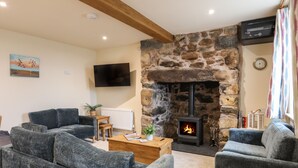 A cozy living room with a stone fireplace, comfortable seating, a flat-screen TV, and a wooden ceiling beam. - Pennant Igyn (Pwllheli)