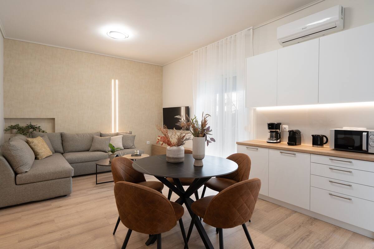 Uma sala de estar moderna com sofá, mesa de centro e poltronas. Há uma área de cozinha com micro-ondas e máquina de café.