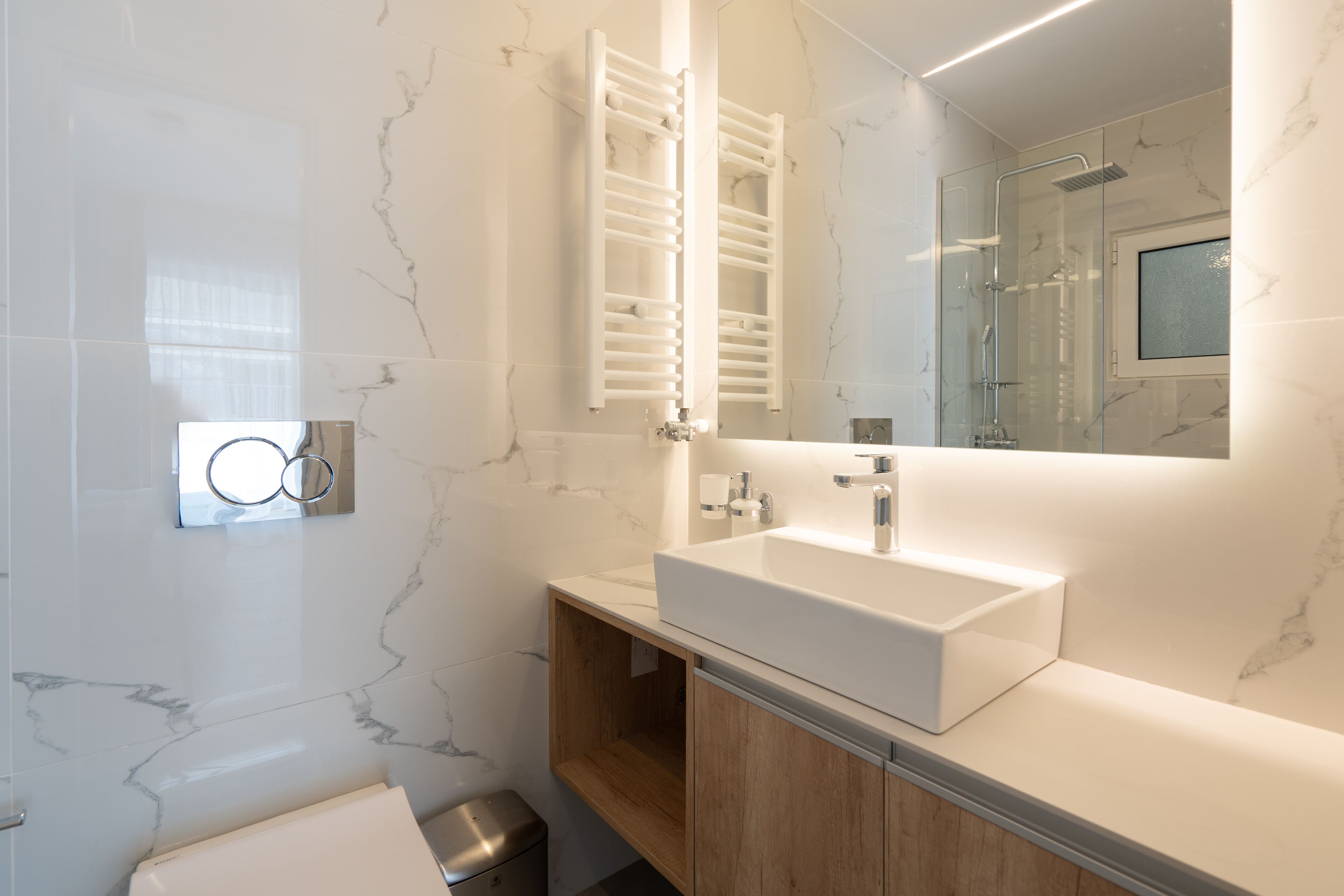 A modern bathroom with a white sink, wooden cabinets, a glass shower enclosure, and a marble-tiled wall.