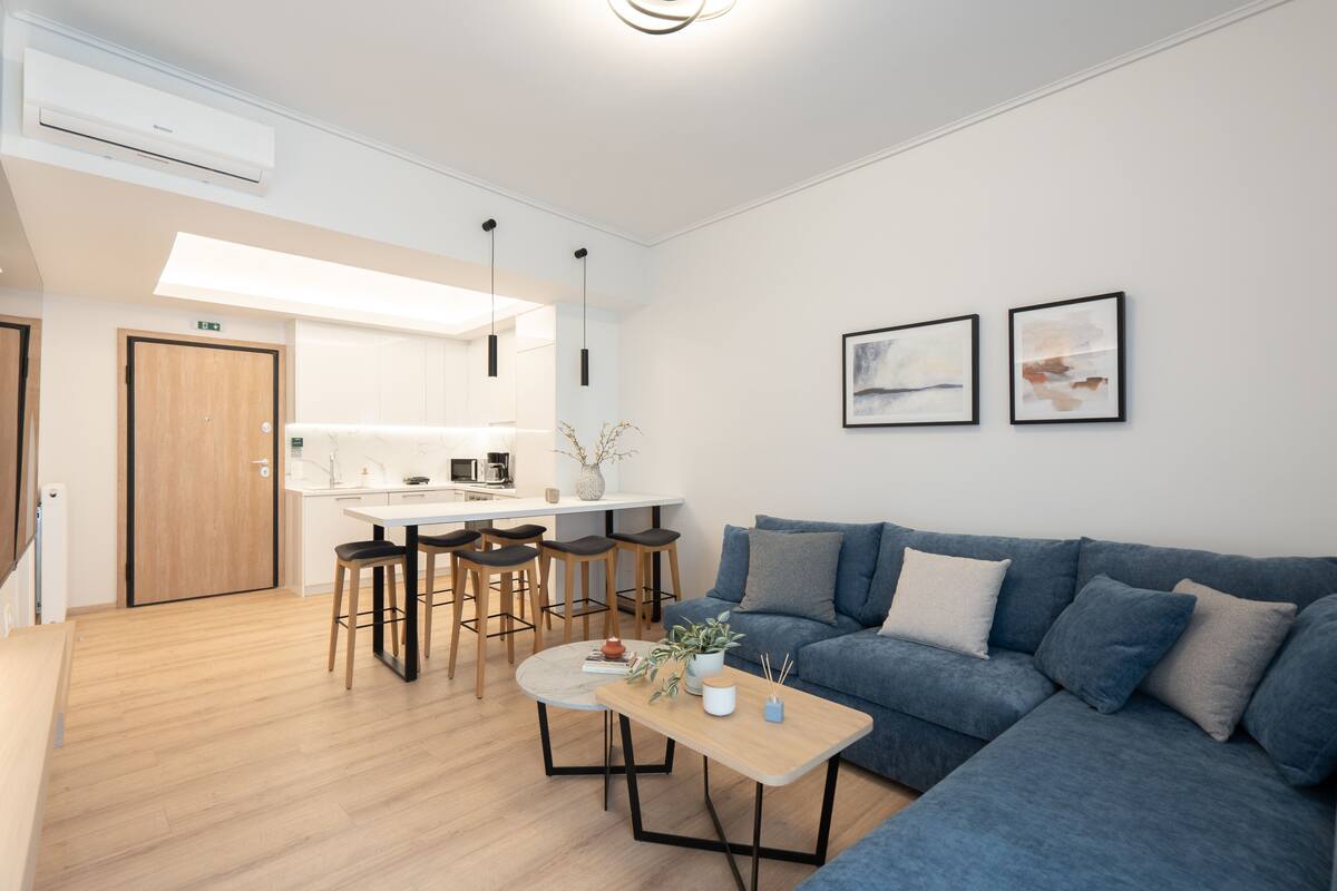 Sala de estar moderna com um sofá azul, uma mesa de centro de madeira e uma área de cozinha com bancada branca e banquetas.