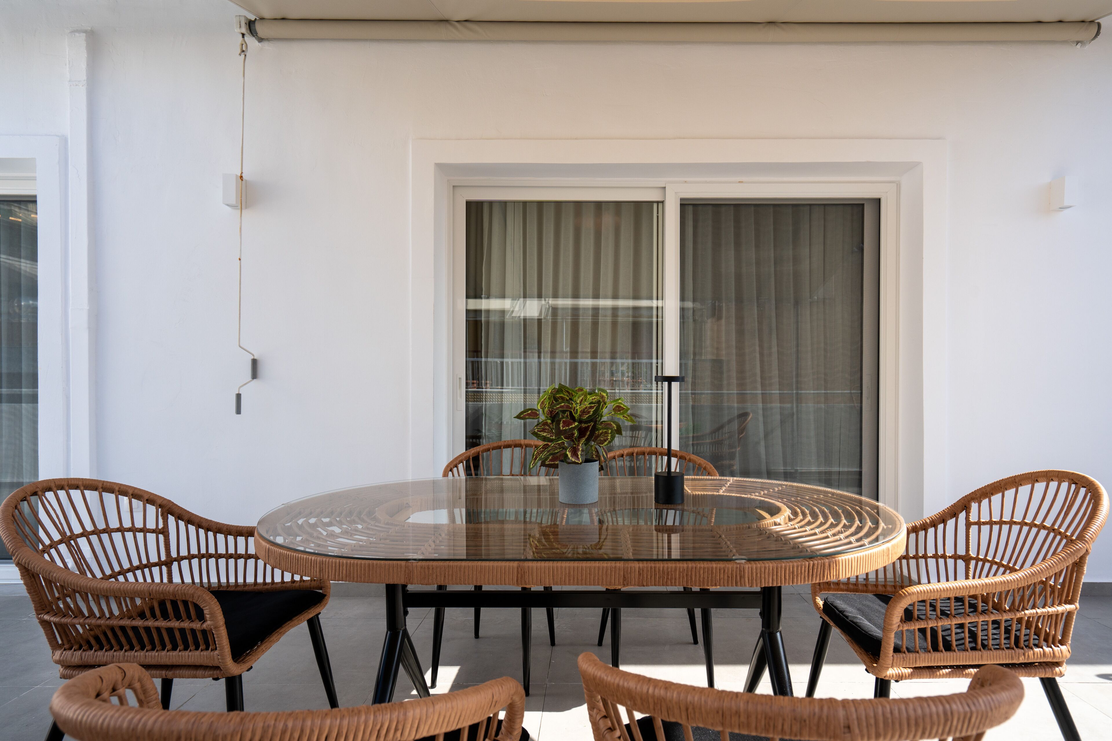 A round glass table with wicker chairs, a potted plant, and a sliding glass door in the background.