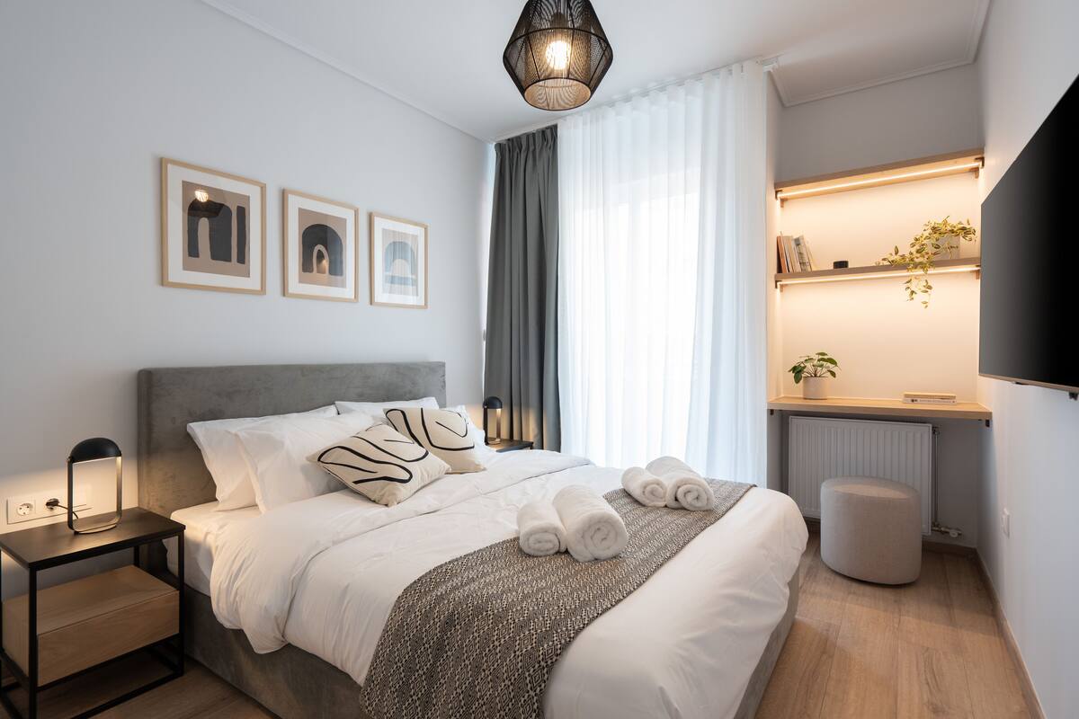 Quarto de hotel moderno com uma cama bem arrumada, luminárias de cabeceira, uma mesinha, um radiador e uma estante com uma planta.