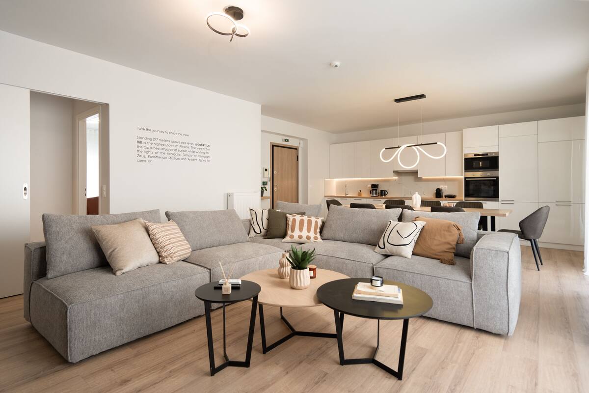 Uma sala de estar moderna com um sofá seccional cinza, duas mesas de centro pretas e uma pequena mesa lateral. Uma cozinha com armários brancos e um forno embutido é visível ao fundo.
