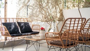 Un espacio de estar al aire libre con muebles de mimbre, cojines y una pequeña mesa, rodeado por una pared blanca y árboles.