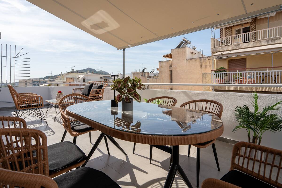 Um terraço no telhado com uma mesa de vidro redonda cercada por cadeiras de vime, uma planta em vaso e vista para a paisagem urbana.