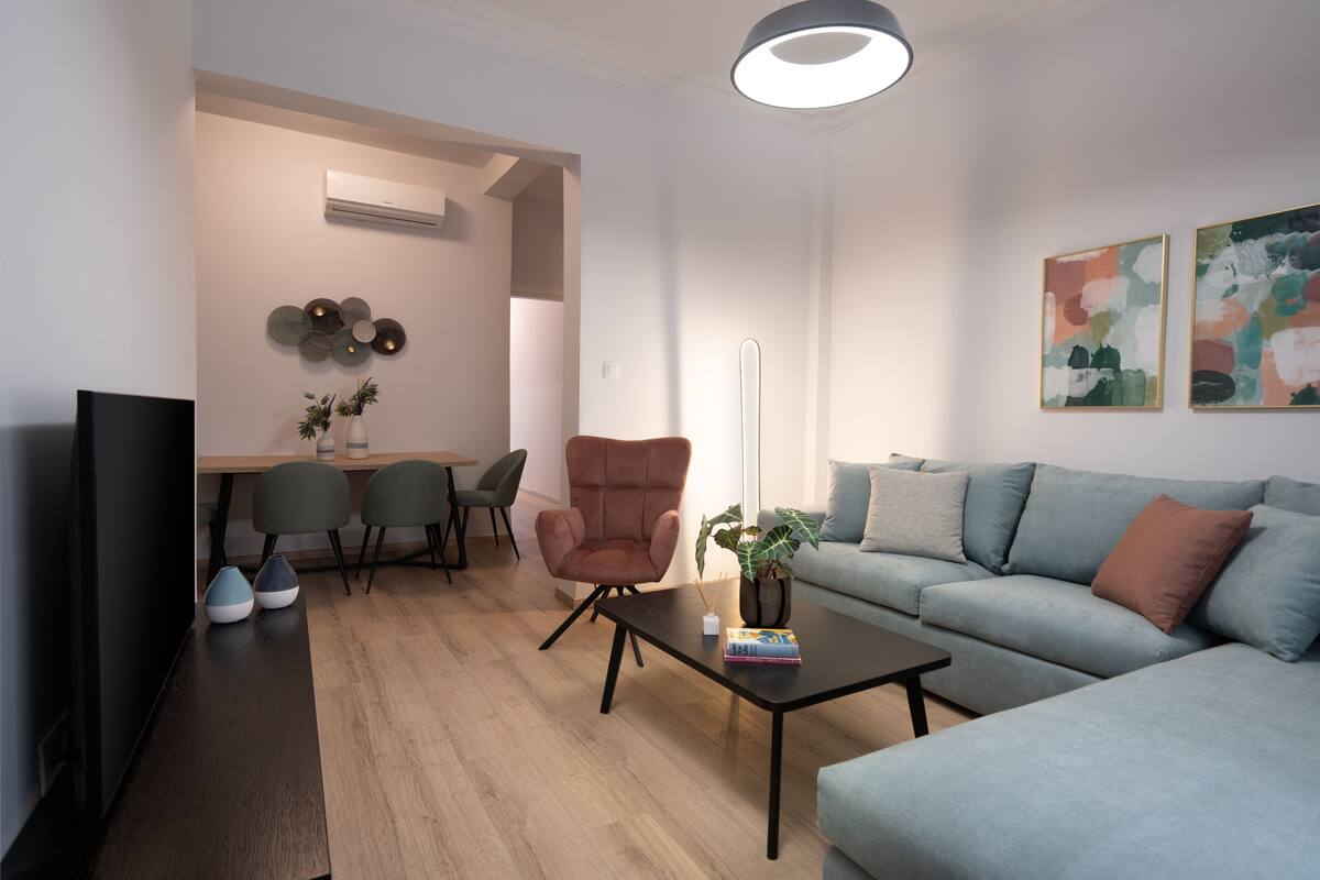 Uma sala de estar moderna com sofá, poltrona, mesa de centro e televisão. Há uma área de jantar com cadeiras e uma mesa, e obras de arte nas paredes.