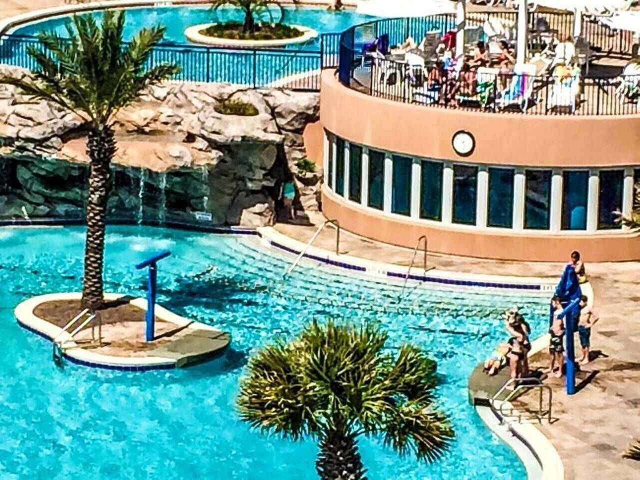 Zona de piscina amb una palmera, una petita illa amb una font i una zona de relax amb gent.