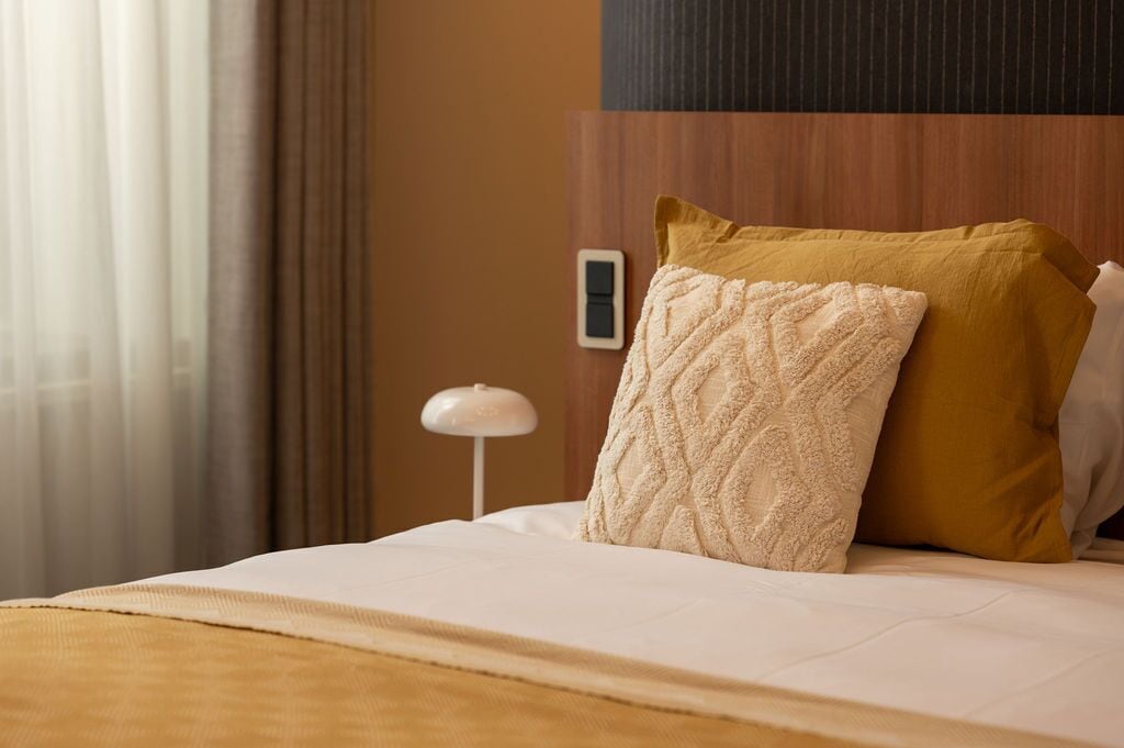 A neatly made bed with a white comforter and two pillows, one with a textured pattern and the other in mustard color, in a room with a wooden headboard and a white nightstand with a lamp.