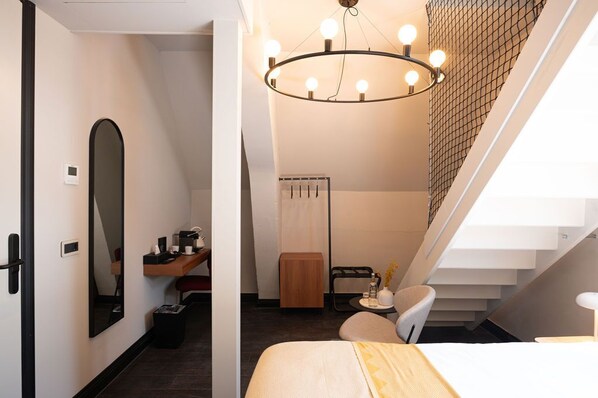 A modern hotel room with a large bed, a desk with a chair, a mirror, a small table with a vase, and a staircase with a railing.