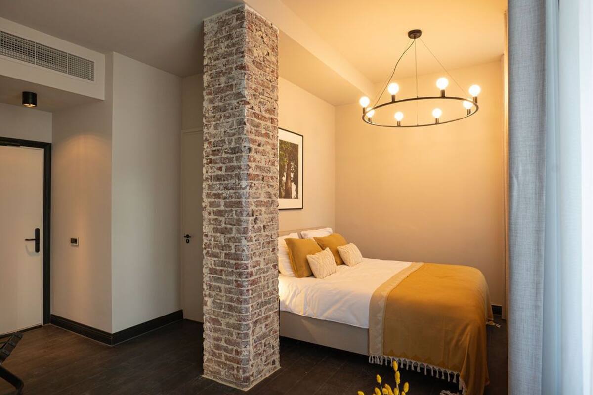 Quarto com um pilar de tijolos, cama com roupa de cama branca, luminária pendente e quadro na parede.