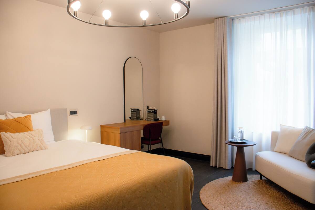 Quarto de hotel com cama, escrivaninha, cadeira, sofá, mesa redonda, espelho e janela com cortinas.