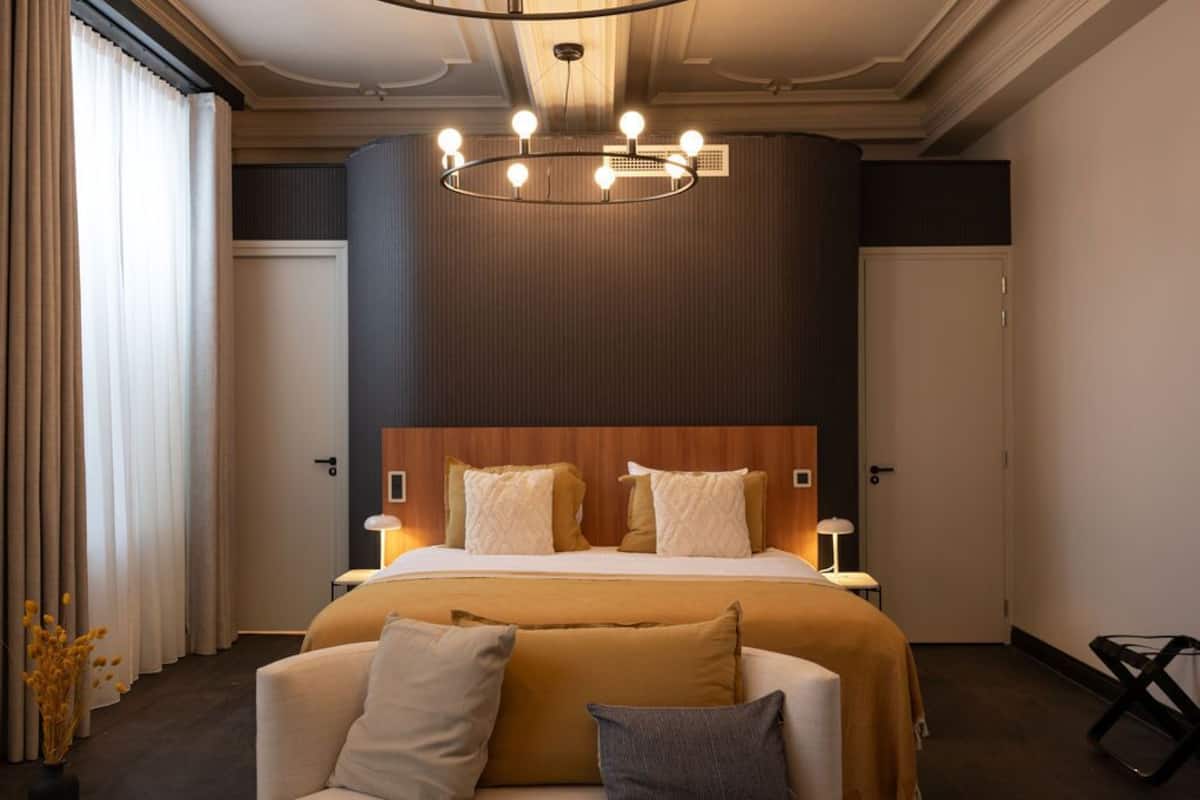 Quarto de hotel com uma cama grande, cabeceira de madeira, duas mesas de cabeceira com luminárias, um sofá e um candelabro.