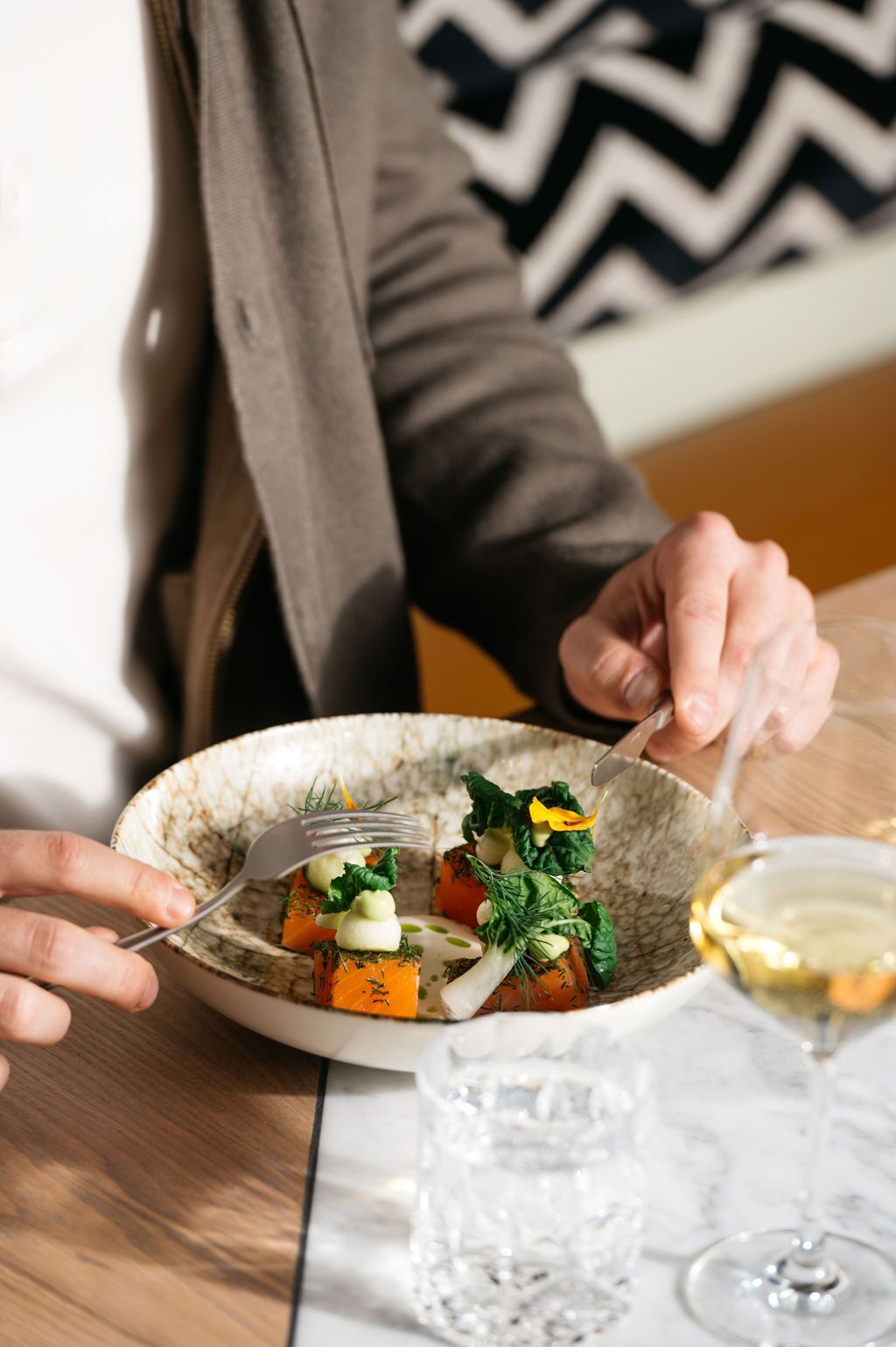 Une personne est assise à une table et prend son repas. Devant elle, il y a un bol en céramique contenant une salade, une fourchette et un verre de vin blanc.