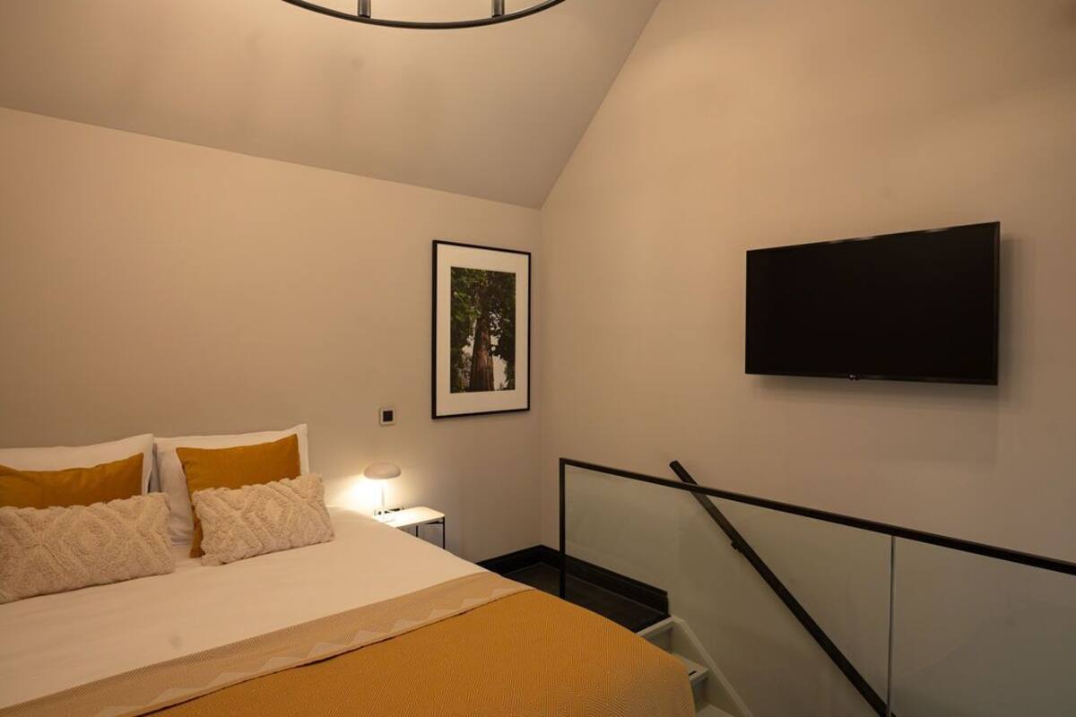 Um quarto com uma cama, um quadro pendurado na parede, uma TV fixada na parede e uma escada com corrimão de vidro.