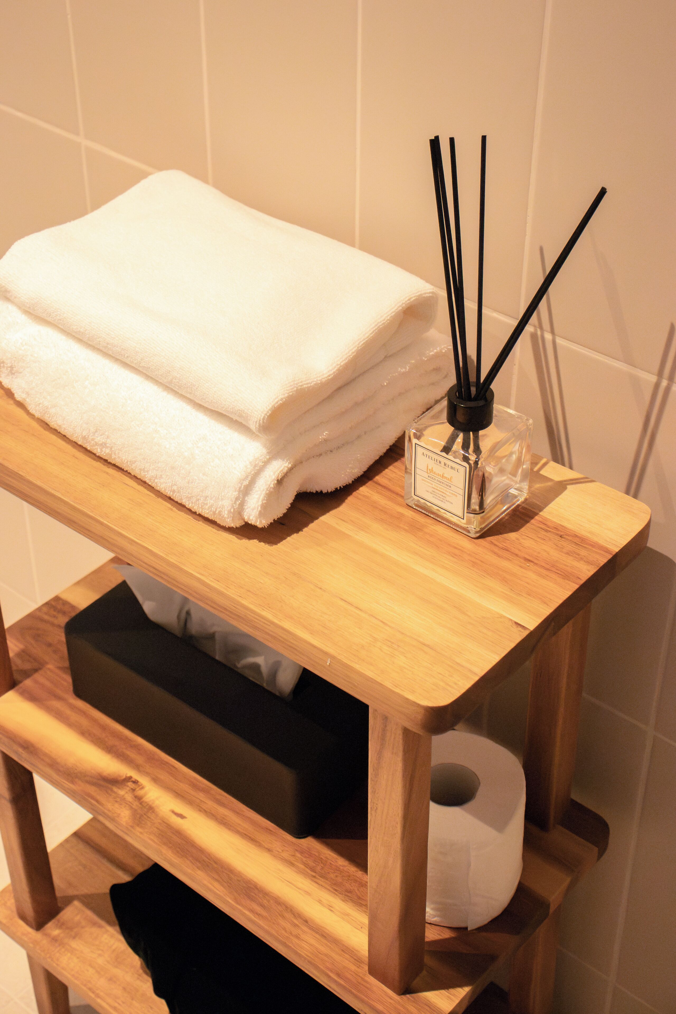 Une étagère en bois sur laquelle est posée une serviette blanche pliée et une bouteille en verre contenant des tiges de roseau.