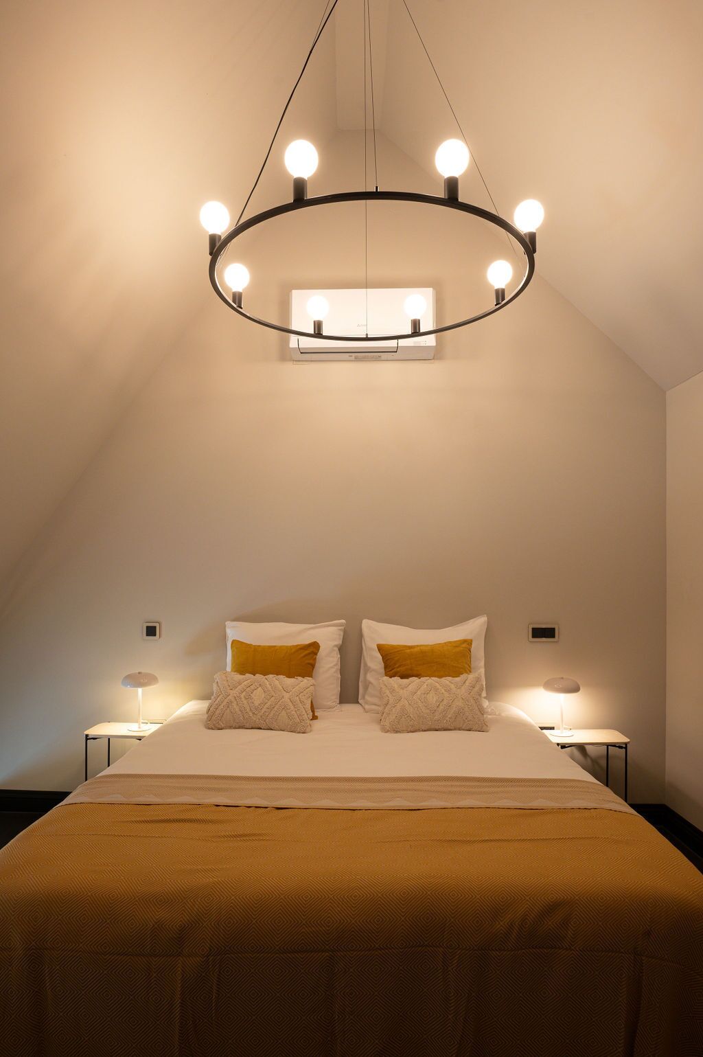 A bedroom with a large bed, two bedside tables, a modern chandelier, and a thermostat on the wall.