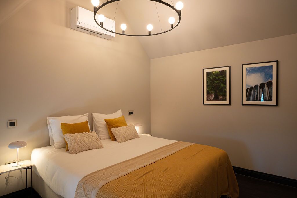 A hotel room with a bed, a bedside table with a lamp, a circular ceiling light, and two framed pictures on the wall.
