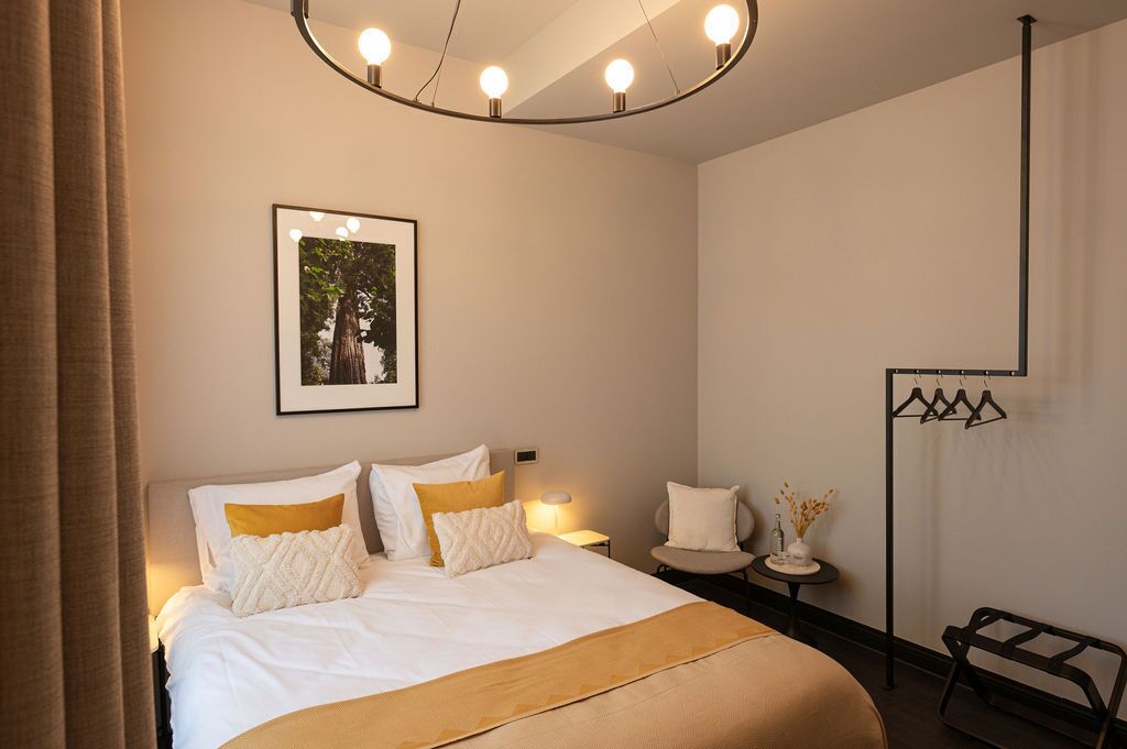 A hotel room with a bed, a bedside table, a chair, a coat rack, and a picture on the wall.