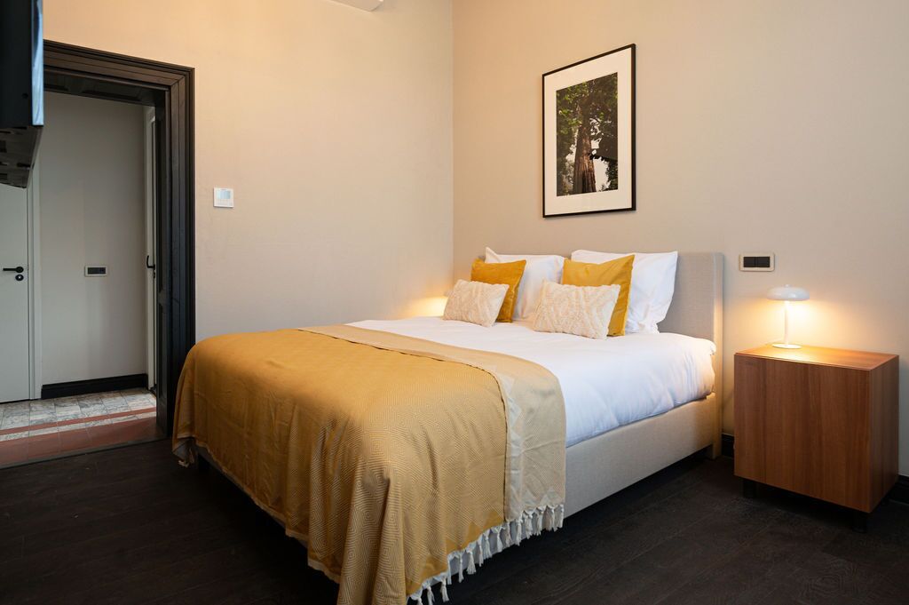 Un dormitorio ordenado con cama, lámparas de noche, un cuadro enmarcado en la pared y una puerta que da a otra habitación.