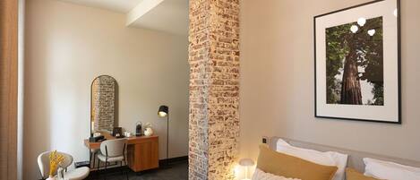 A hotel room with a bed, a desk, and a chair. There is a brick wall and a framed picture on the wall.