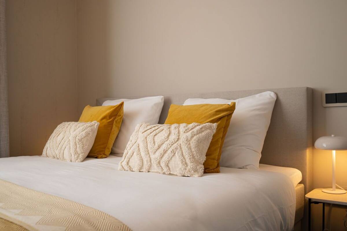 Cama bem arrumada com almofadas brancas e mostarda, abajur e manta texturizada.