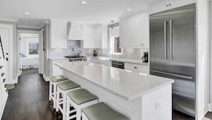 A modern kitchen with white cabinetry, a large stainless steel refrigerator, and a central island with bar stools. - Ocean 180 | True Oceanfront End Unit with 180° Views in Wild Dunes (Isle of Palms)
