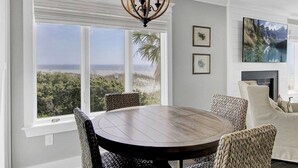 A dining area with a round table and chairs, large windows with a view of the beach, a chandelier, and framed artwork on the wall. - Ocean 180 | True Oceanfront End Unit with 180° Views in Wild Dunes (Isle of Palms)