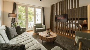 Ett modernt vardagsrum med en soffa, en fåtölj och ett runt soffbord. En tv är monterad på väggen, och det finns ett stort fönster med utsikt över byggnaderna.