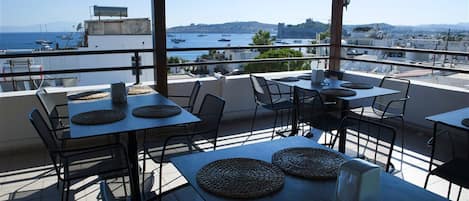 An outdoor terrace with tables and chairs, overlooking a marina with boats and a distant view of hills.