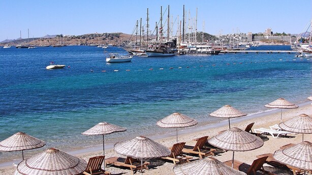 A beach with sun loungers and thatched umbrellas, a marina with sailboats, and a hilly town in the background. - Smart City Suites Bodrum (Bodrum)
