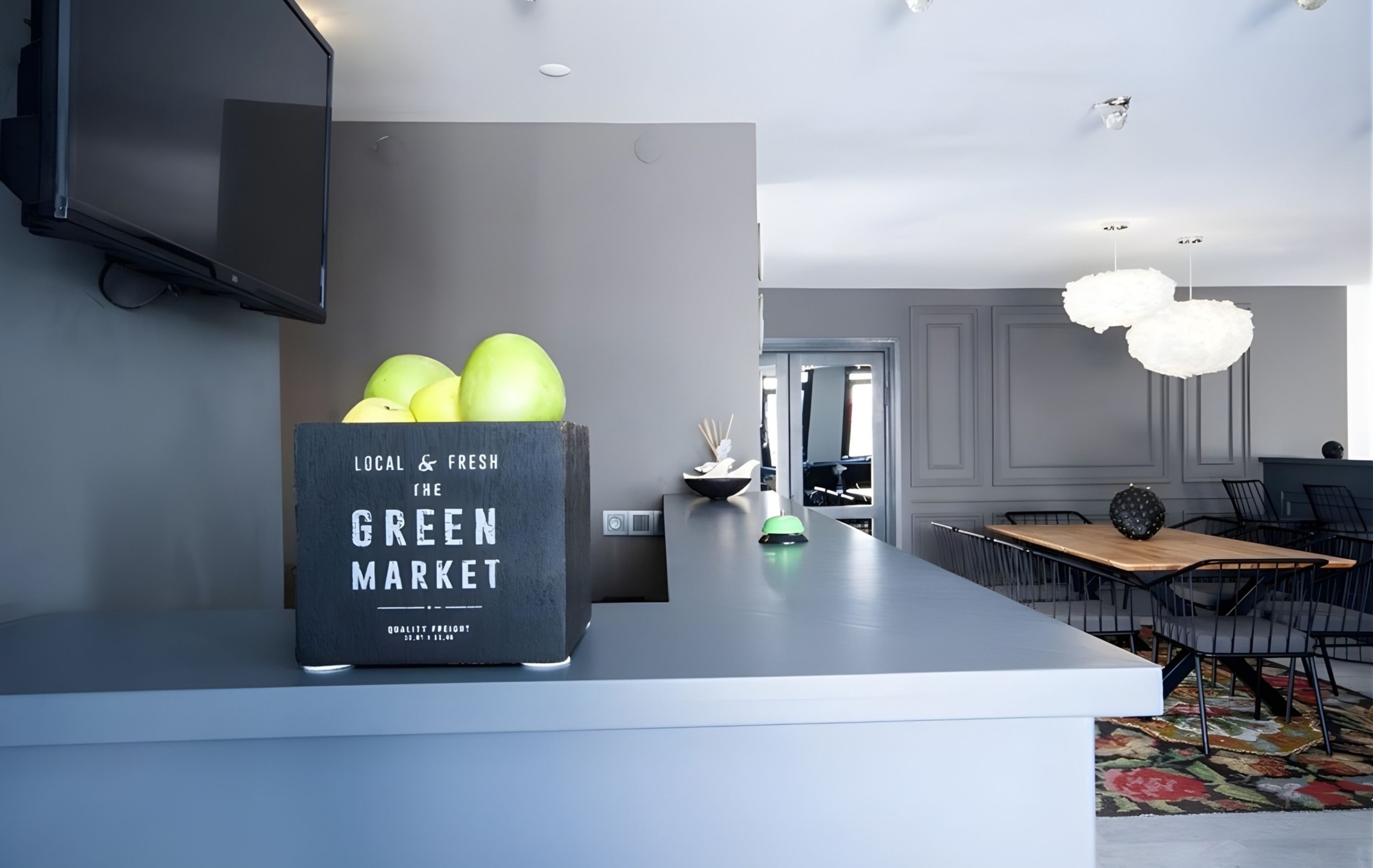 Una cocina moderna con encimera gris, una caja negra con la etiqueta 'Local & Fresh The Green Market' y una zona de comedor con mesa y sillas de madera.