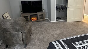 Ein Wohnzimmer mit Fernseher, Kamin, grauem Sofa und einem Kleiderschrank mit Kleidung.