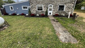 Ein zweistöckiges Haus mit Steinaußenwand, einer Veranda und einem kleinen Garten mit roten und weißen Blumen.