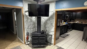 Ein Wohnzimmer mit Kamin, einem an der Wand montierten Fernseher und einer durch eine offene Tür einsehbaren Küchenzeile.