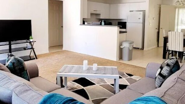 Una sala moderna con un sofá gris, una mesa de centro y un televisor. Se observa una zona de cocina con armarios blancos y un refrigerador.