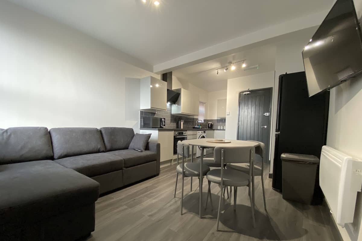 A modern living room with a grey sectional sofa, a dining table with four chairs, a kitchen area with a microwave, oven, and sink, and a grey door.