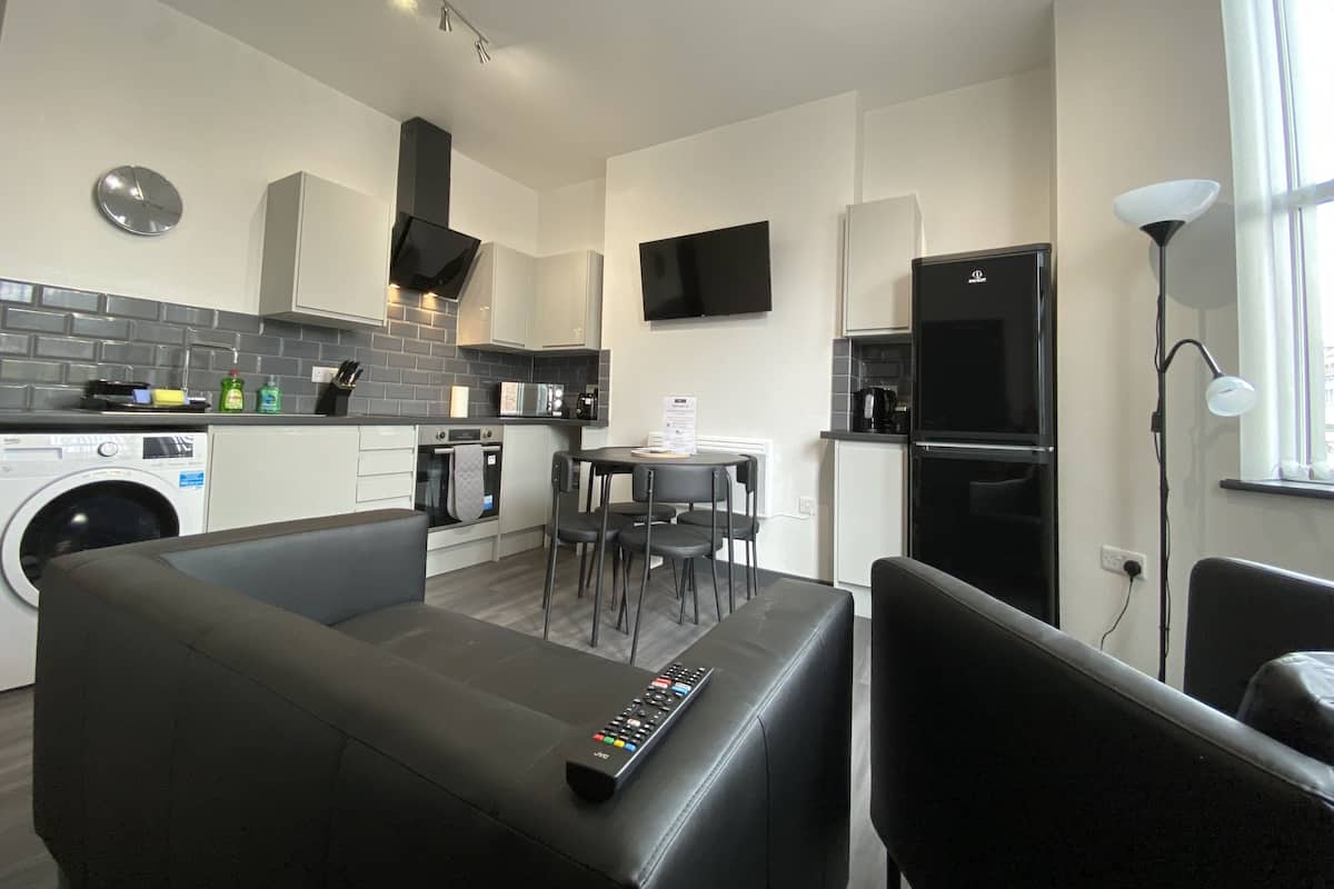 A modern kitchen with a washing machine, a dining area with a table and chairs, and a black sofa with a remote control.