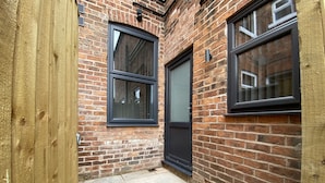 A brick building with two windows and a door, a stone-paved courtyard, and a wooden fence. - StayCrewe Apartments (Crewe)