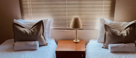 A bedroom with two beds, a nightstand, a lamp, and a window with blinds.