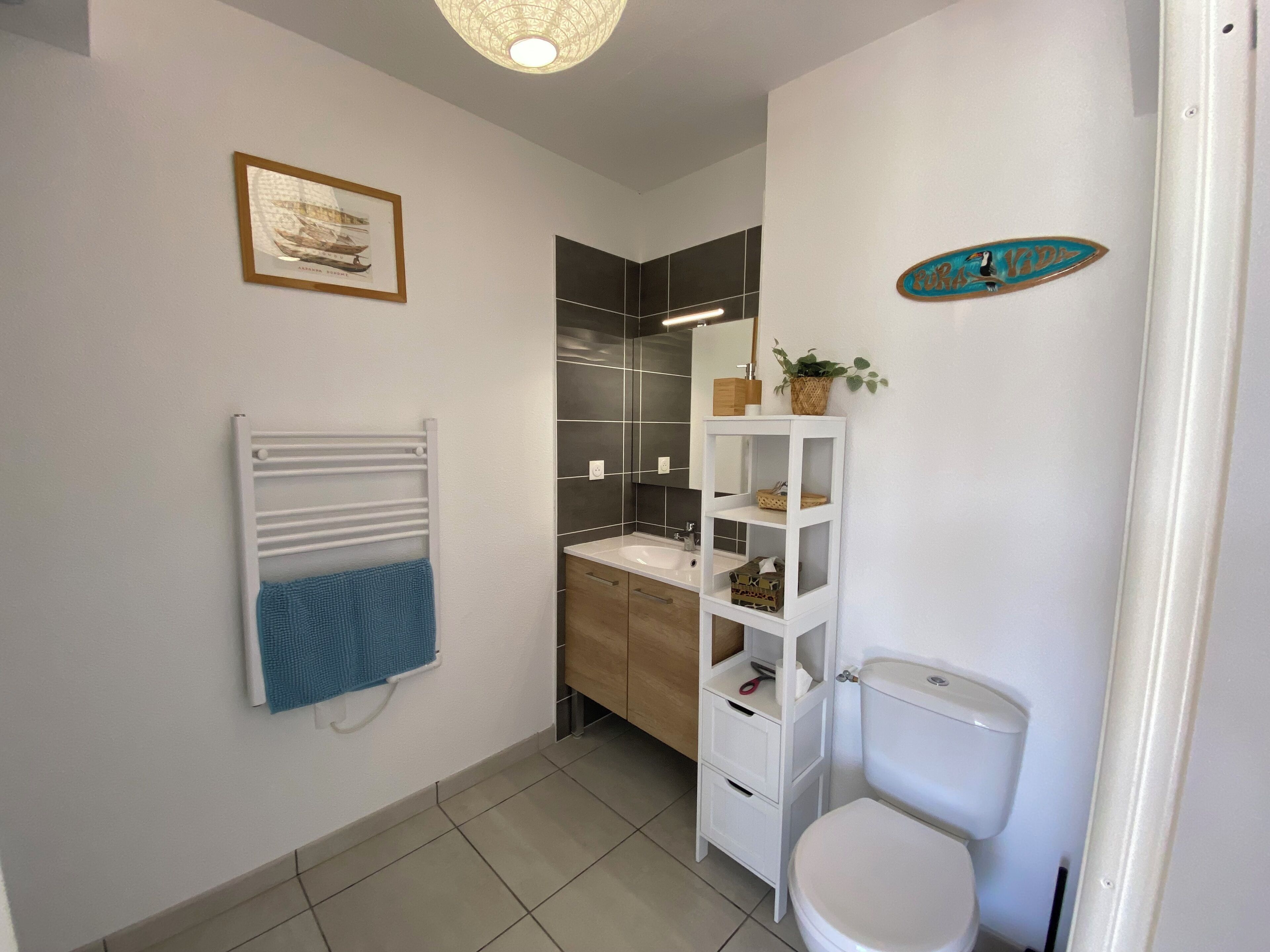 A modern bathroom with a white sink, a toilet, a towel rack with a blue towel, a framed picture on the wall, and a shelf with toiletries.