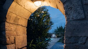 Un arc en pierre surmonté d’un luminaire, qui mène à un sentier recouvert de gravier et bordé de verdure.