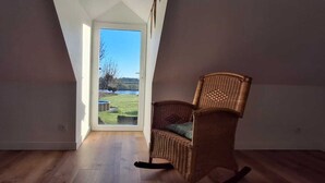 Une pièce avec un plafond haut, un fauteuil en osier et une vue sur un espace verdoyant et un plan d’eau, le tout visible à travers une grande fenêtre.